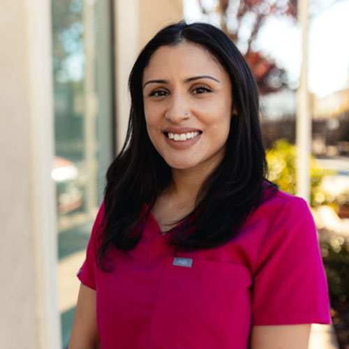 A woman with long dark hair wearing a magenta scrub top stands outside, smiling at the camera, with sunlight and blurred greenery in the background.