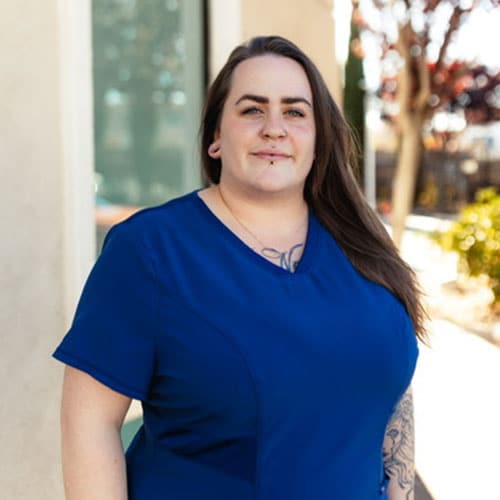 A woman with long brown hair, wearing a royal blue top, stands outside near a building. She has tattoos on her chest and arm, and is looking at the camera with a neutral expression. Trees and sunlight are in the background.